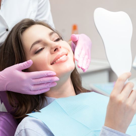 Woman in dental chair looking at her reflection in a tooth-shaped hand mirror